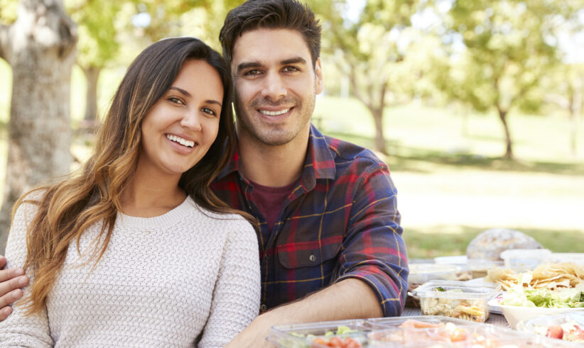 A smiling couple sits close together at a picnic table outdoors, surrounded by various food containers, with trees and greenery in the background.