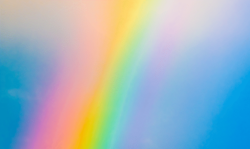 A vibrant rainbow with distinct colors arcs across a soft blue sky, blending into light, wispy clouds. The colors are bright and vivid, creating a cheerful and uplifting atmosphere.