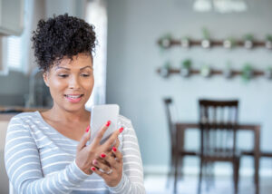 A woman with curly hair and red nails, wearing a gray and white striped shirt, smiles while looking at her smartphone in a bright kitchen or dining area with plants on the wall in the background.
