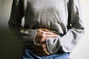 A person wearing a gray long-sleeve shirt and blue jeans holds their hands over their stomach, suggesting discomfort or pain.