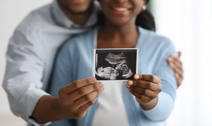A smiling couple embraces and holds up an ultrasound image, suggesting excitement and anticipation for their expected baby. Their faces are partially out of focus, emphasizing the ultrasound photo.
