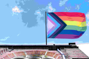 A Progress Pride flag waves atop a brick building against a blue sky with clouds. The flag features rainbow stripes and chevrons in black, brown, light blue, pink, and white.