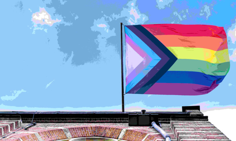A Progress Pride flag waves atop a brick building against a blue sky with clouds. The flag features rainbow stripes and chevrons in black, brown, light blue, pink, and white.