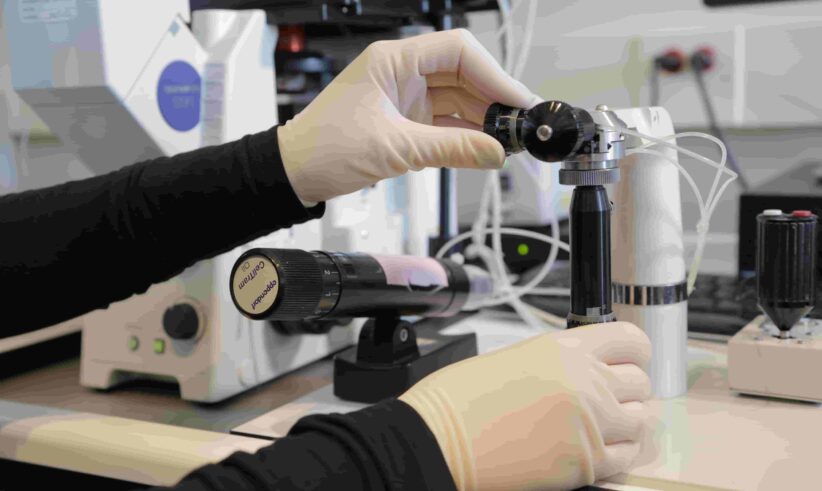 A person wearing gloves adjusts scientific equipment in a laboratory setting, surrounded by various instruments and cables on a workbench.