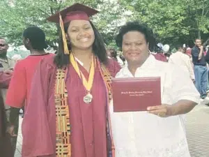 A student in a maroon graduation cap and gown stands smiling next to a woman in white holding a diploma. They are outdoors with other people and trees in the background.