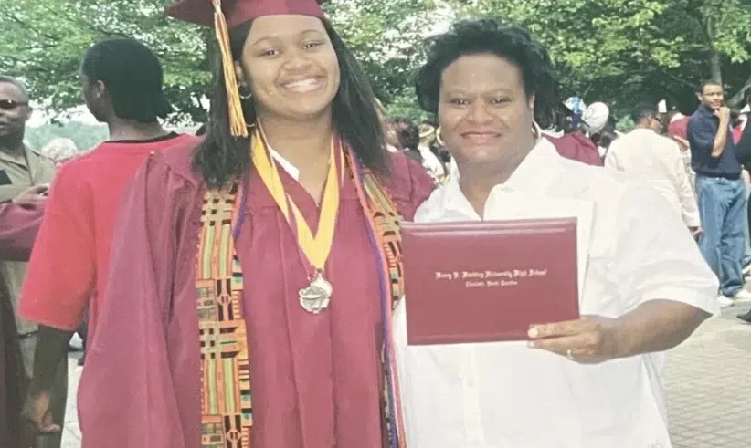 A student in a maroon graduation cap and gown stands smiling next to a woman in white holding a diploma. They are outdoors with other people and trees in the background.