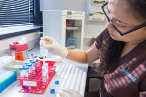 A woman wearing gloves uses a pipette to transfer liquid into a test tube in a laboratory. Various lab equipment, test tubes, and a biohazard container are visible on the counter.