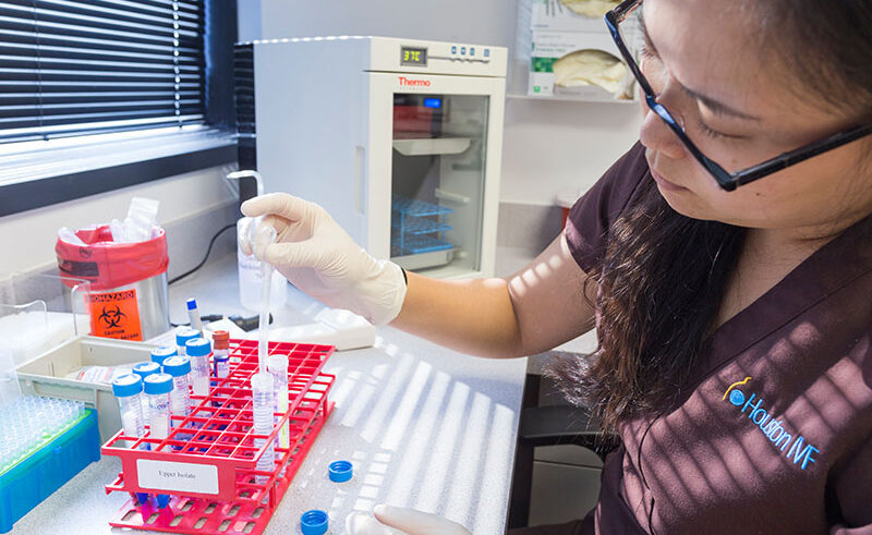 A woman wearing gloves uses a pipette to transfer liquid into a test tube in a laboratory. Various lab equipment, test tubes, and a biohazard container are visible on the counter.