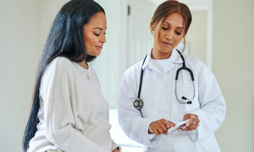 A doctor in a white coat and stethoscope discusses information on a tablet with a smiling woman who is sitting on an exam table. Both appear focused and engaged in conversation in a bright medical office.