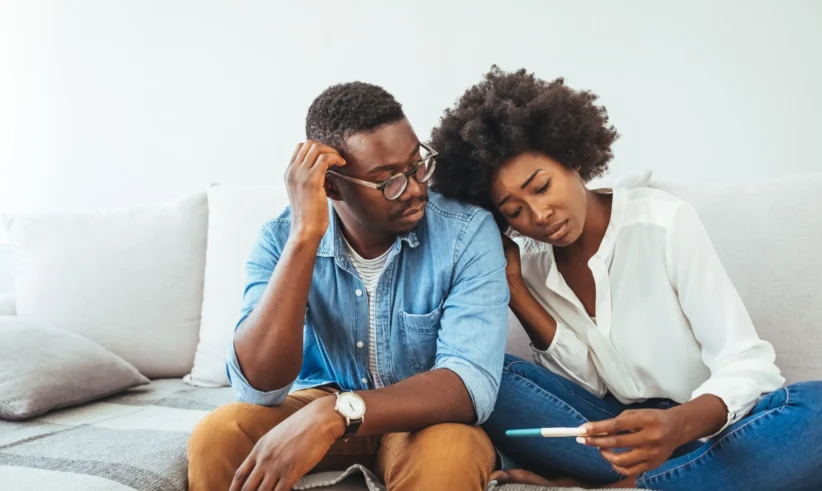 A couple sits together on a couch, looking concerned while the woman holds a pregnancy test. The man touches his head with worry, and the woman leans on his shoulder, appearing anxious and thoughtful.