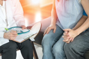 A doctor holding a clipboard and pen speaks with a couple sitting closely together, holding hands, suggesting a medical consultation or discussion about health.