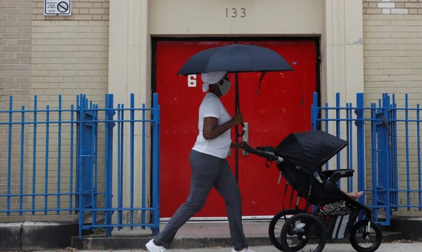 A person wearing a mask walks past a red door at Public School 133, pushing a stroller and holding a black umbrella. Blue metal fencing lines the sidewalk.