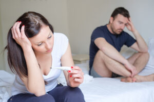 A woman sits on a bed holding a thermometer, looking worried, while a man sits behind her, appearing concerned and pensive. Both have serious expressions in a bright, neutral-colored room.
