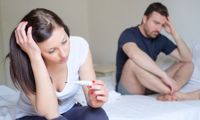 A woman sits on a bed holding a thermometer, looking worried, while a man sits behind her, appearing concerned and pensive. Both have serious expressions in a bright, neutral-colored room.