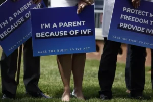 Three people stand on grass holding blue signs supporting IVF; the center sign reads IM A PARENT BECAUSE OF IVF and the others have similar messages. Their faces are not visible.