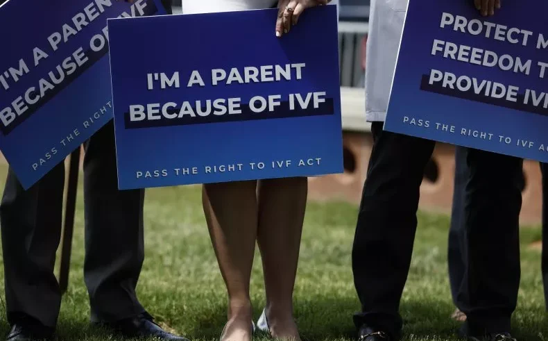 Three people stand on grass holding blue signs supporting IVF; the center sign reads IM A PARENT BECAUSE OF IVF and the others have similar messages. Their faces are not visible.