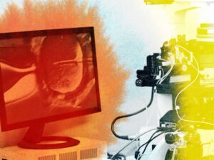 A computer monitor displays a magnified image of a cell, while scientific equipment, including a microscope, is set up beside it, bathed in red and yellow lighting effects.