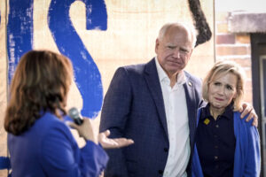 A woman holding a microphone speaks to a man and woman standing together. The man has his arm around the woman, and both are wearing blue jackets. A large sign with the letters IS is visible in the background.