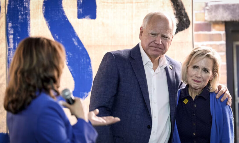 A woman holding a microphone speaks to a man and woman standing together. The man has his arm around the woman, and both are wearing blue jackets. A large sign with the letters IS is visible in the background.
