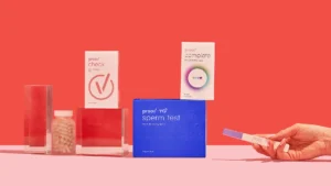 A hand holds a pregnancy test beside various fertility and health products, including Proov check, Proov complete, Proov sperm test, and a bottle of capsules, all arranged on a pink surface with a red background.