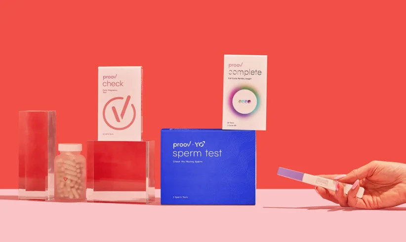 A hand holds a pregnancy test beside various fertility and health products, including Proov check, Proov complete, Proov sperm test, and a bottle of capsules, all arranged on a pink surface with a red background.