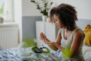 A woman with curly hair sits on a bed, using a blood glucose meter to check her blood sugar. She holds a test strip to her finger, with diabetes supplies on a tray in front of her.