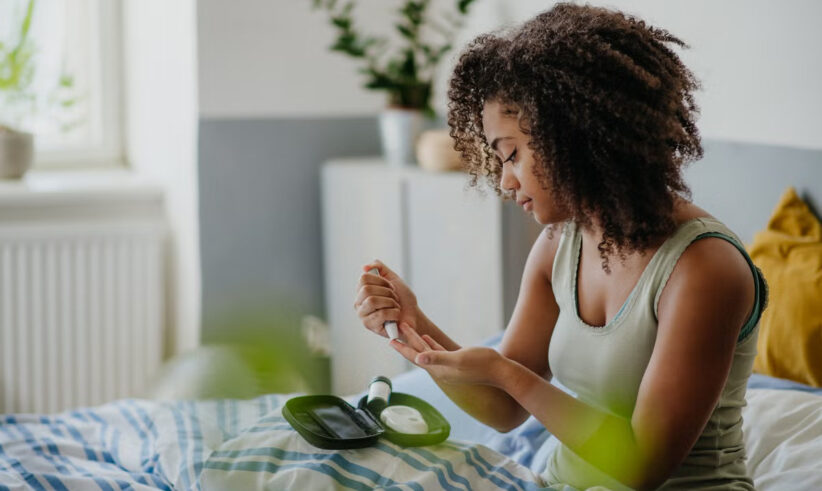 A woman with curly hair sits on a bed, using a blood glucose meter to check her blood sugar. She holds a test strip to her finger, with diabetes supplies on a tray in front of her.