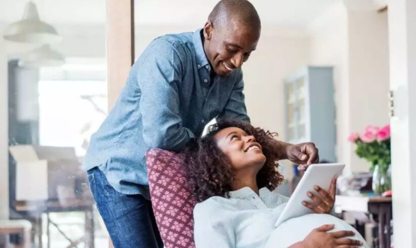 A smiling man leans over a seated pregnant woman who is holding a tablet. They are indoors, looking at each other affectionately, with the woman appearing relaxed and comfortable. Flowers and household items are visible in the background.