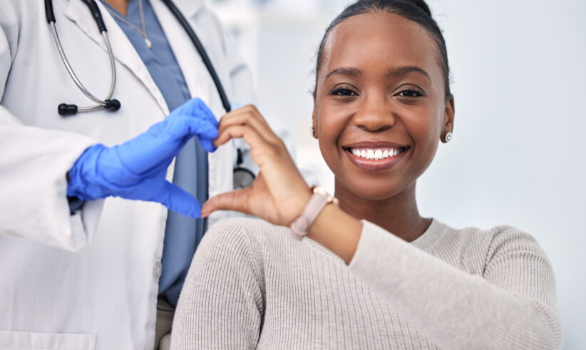A smiling woman and a doctor wearing a white coat and blue gloves form a heart shape with their hands, symbolizing care and support, in a bright medical setting.