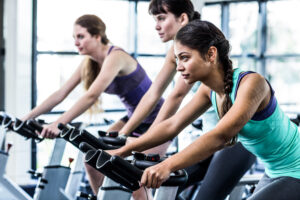 Three women in athletic wear ride stationary bikes in a bright gym, focused and determined, exercising together in a row.
