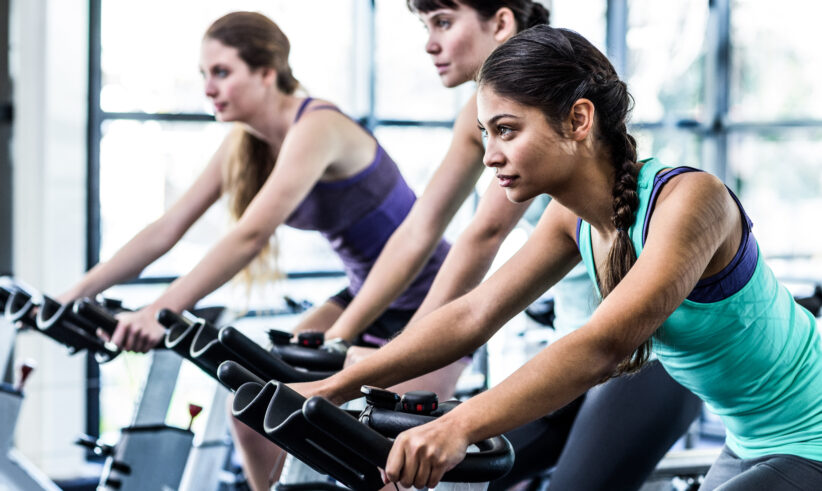 Three women in athletic wear ride stationary bikes in a bright gym, focused and determined, exercising together in a row.
