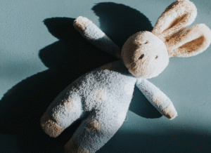 A plush bunny toy with long ears lies on a blue surface, casting a shadow to the left. The bunny appears soft and slightly worn, with light cream and blue coloring.