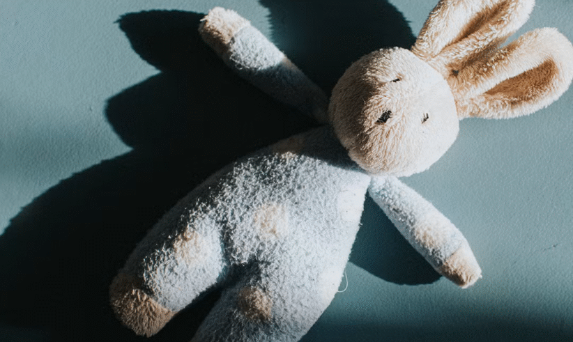 A plush bunny toy with long ears lies on a blue surface, casting a shadow to the left. The bunny appears soft and slightly worn, with light cream and blue coloring.