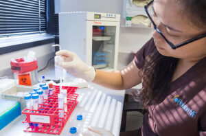 A scientist wearing gloves and a lab coat uses a pipette to transfer liquid into test tubes organized in a red rack on a laboratory bench, with lab equipment and a fridge in the background.