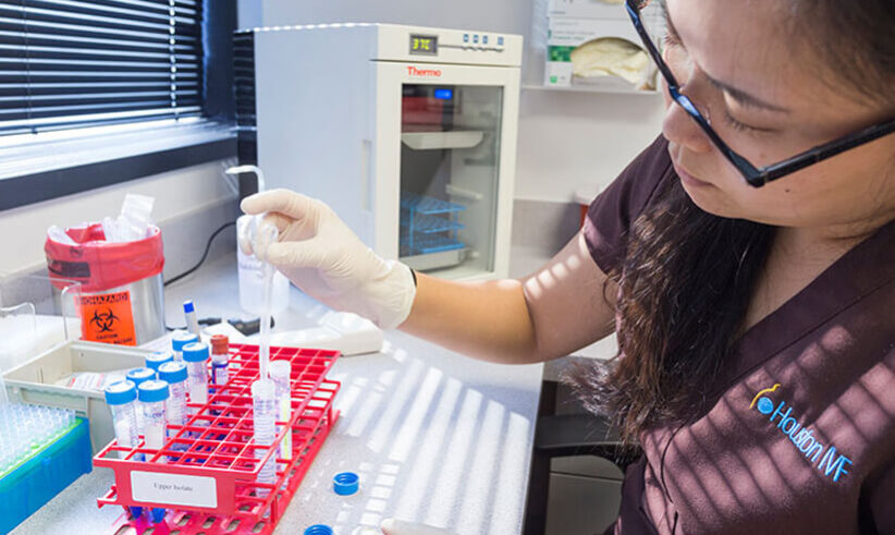A scientist wearing gloves and a lab coat uses a pipette to transfer liquid into test tubes organized in a red rack on a laboratory bench, with lab equipment and a fridge in the background.
