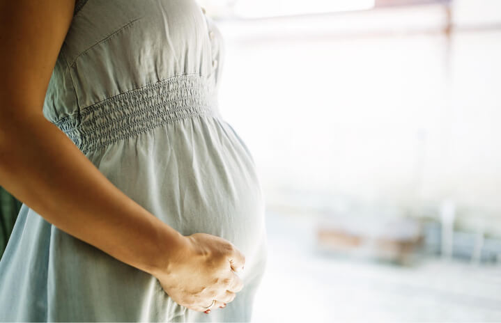A close-up of a pregnant woman wearing a light gray dress, gently holding her belly with one hand, standing indoors with a soft background.