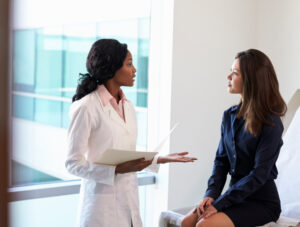 A female doctor in a white coat holding a clipboard talks to a woman sitting on an exam table in a medical office, both appearing engaged in conversation.
