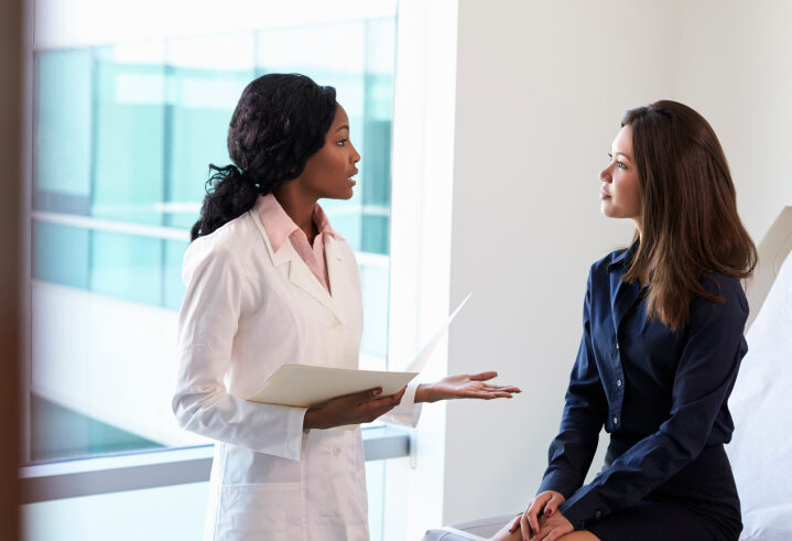 A female doctor in a white coat holding a clipboard talks to a woman sitting on an exam table in a medical office, both appearing engaged in conversation.