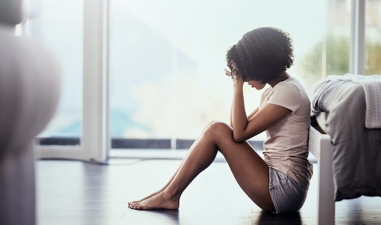 A young woman sits on the floor beside a bed with her head in her hands, appearing stressed or upset, in a bright room with large windows in the background.