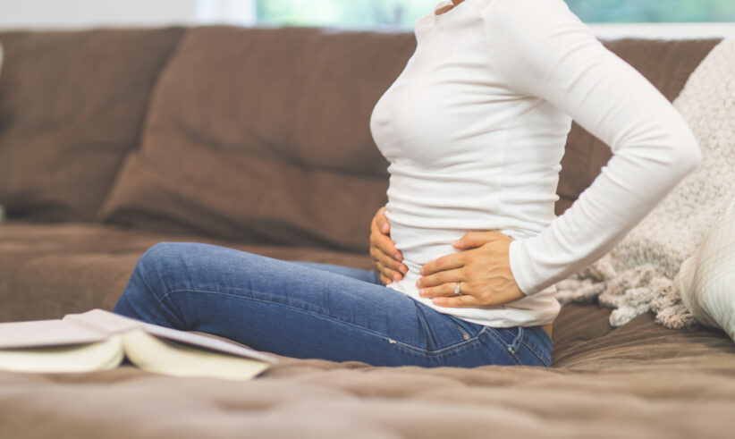 A woman sits on a couch holding her stomach with both hands, possibly experiencing abdominal discomfort. An open book lies next to her on the brown sofa.