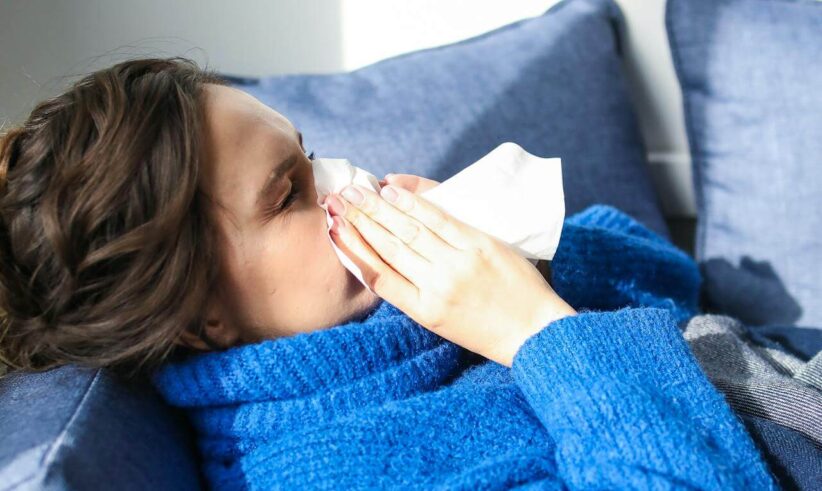 A person wearing a blue sweater sits on a couch and holds a tissue to their nose, appearing to sneeze or blow their nose. Blue cushions are in the background, and sunlight comes through a nearby window.