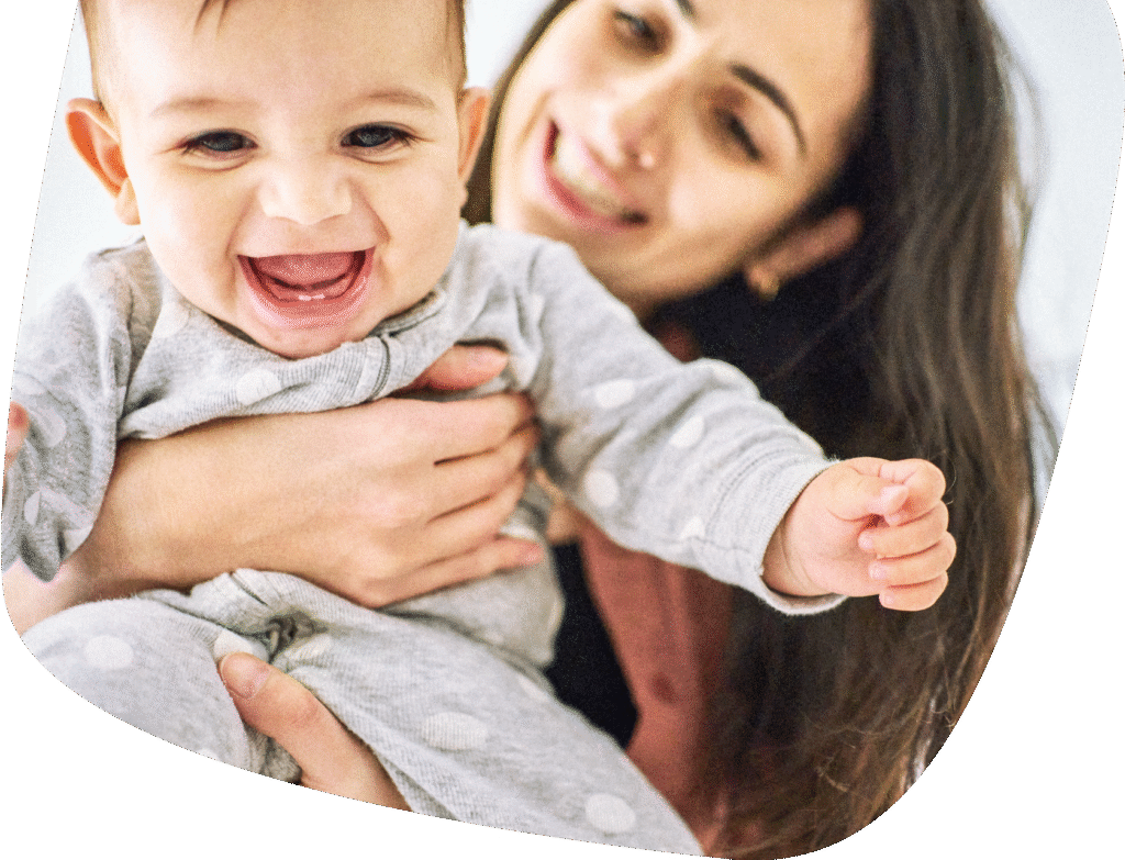 A smiling baby in a grey polka-dot onesie is held by a woman with long dark hair, who is also smiling and looking at the baby. The scene conveys warmth and happiness.
