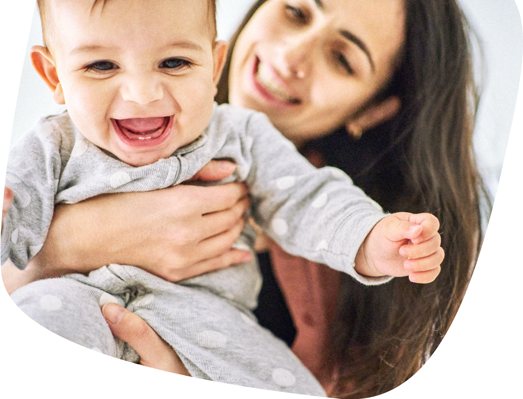 A smiling baby in a grey polka-dot onesie is held by a woman with long dark hair, who is also smiling and looking at the baby. The scene conveys warmth and happiness.