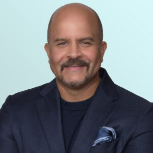 A smiling man with a mustache and beard, wearing a dark blue blazer, matching sweater, and a blue pocket square, poses in front of a light blue background.