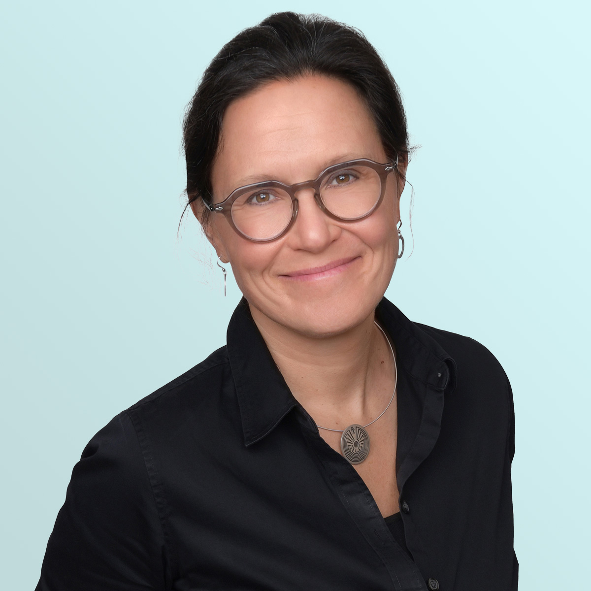 A woman with dark hair pulled back, wearing glasses, a black collared shirt, and a round pendant necklace, smiles at the camera against a light blue background.