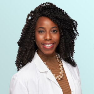 A smiling woman with curly hair wearing a white lab coat, a brown turtleneck, and a pearl necklace, posing in front of a light blue background.
