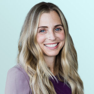 A woman with long, wavy blonde hair smiles at the camera. She is wearing a mauve jacket over a purple top and stands in front of a light blue background.