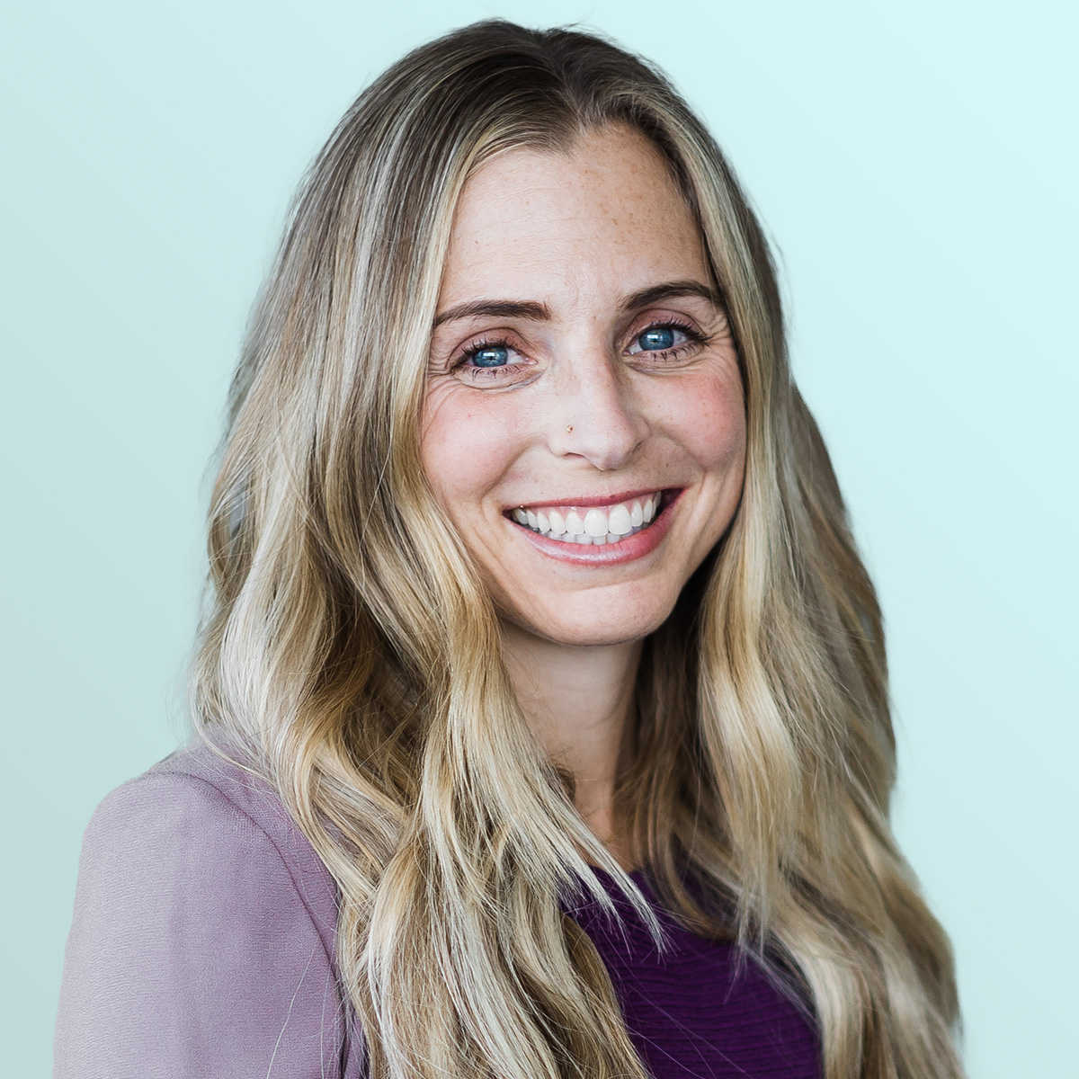 A woman with long, wavy blonde hair smiles at the camera. She is wearing a mauve jacket over a purple top and stands in front of a light blue background.