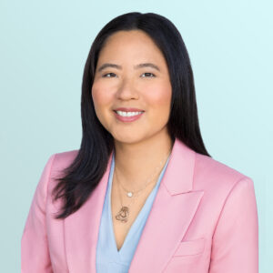 A woman with long dark hair wearing a light pink blazer and light blue blouse, smiling at the camera. She has layered necklaces and is posed against a pale blue background.