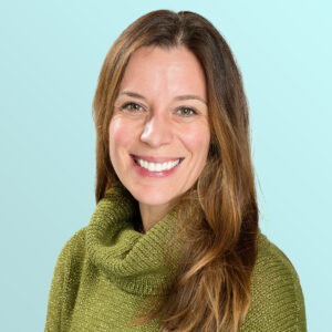 A woman with long brown hair, wearing a green turtleneck sweater, smiling against a light blue background.